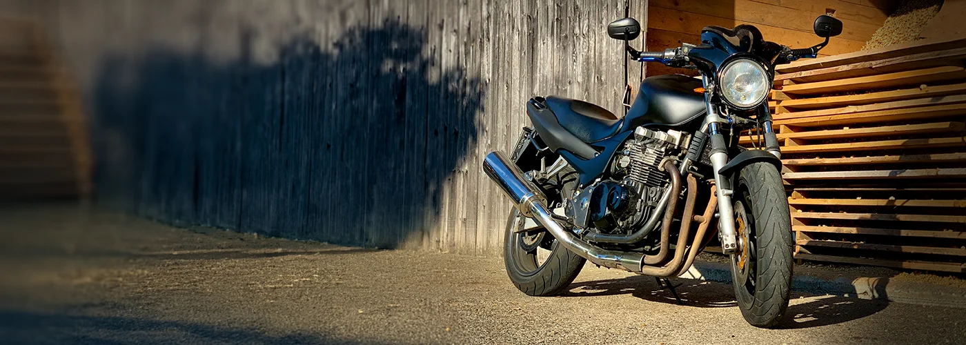 Schwarze Kawasaki in Dreiviertelansicht von vorne, geparkt auf Asphalt vor einer verwitterten Holzwand und gestapelten Holzlatten. Das Motorrad verfügt über einen runden Frontscheinwerfer, einen freiliegenden Reihenmotor mit vier Krümmern, einen silbernen Auspuff und breite Straßenreifen