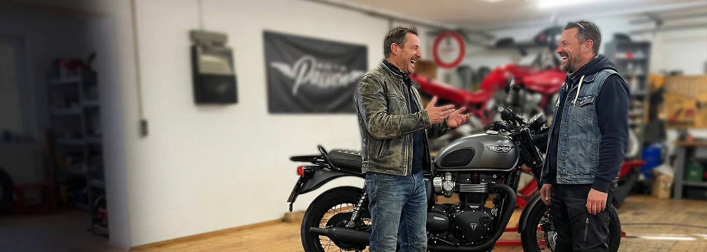 Zwei Motorrad-Enthusiasten fachsimpeln in der Werkstatt von MotoPassion über eine Triumph Modern Classic. Die Szene vermittelt persönliche Beratung, technische Expertise und Leidenschaft für Motorräder.