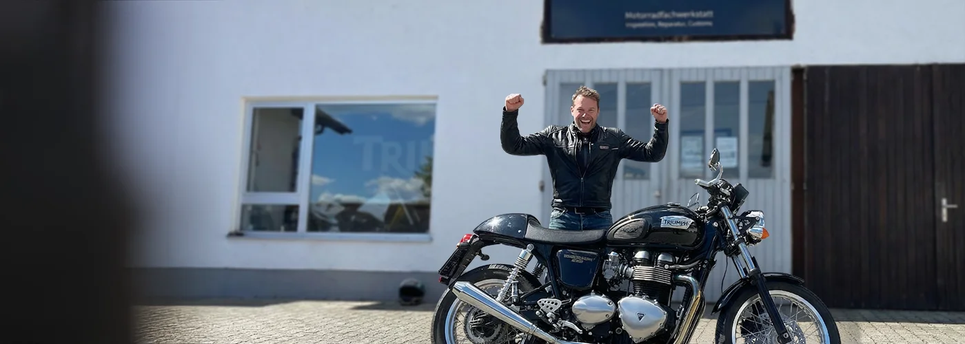 Glücklicher Kunde mit erhobenen Armen neben einer Triumph Thruxton Café Racer vor unserer Motorrad&shy;werkstatt in Emmering bei München.